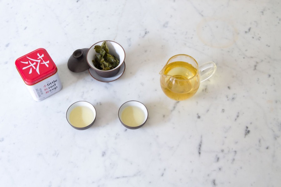 A tin of Little Red Cup tea beside a gaiwan of wet oolong leaf, a glass pitcher of pale gold tea, and two cups.