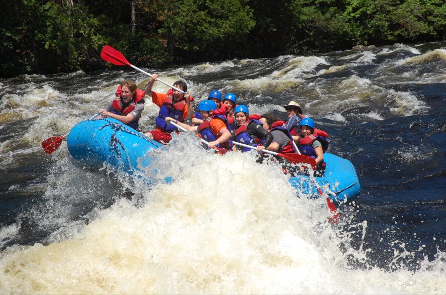 Eight people in helmets and life jackets paddling a blue raft through a large standing wave on a fast river.